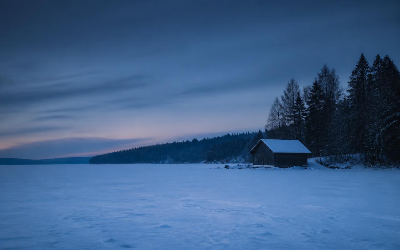 Zugefrorener, schneebedeckter See in blauer Dämmerung, am Ufer eine einzelne dunkle Bootshütte vor Nadelwald.
